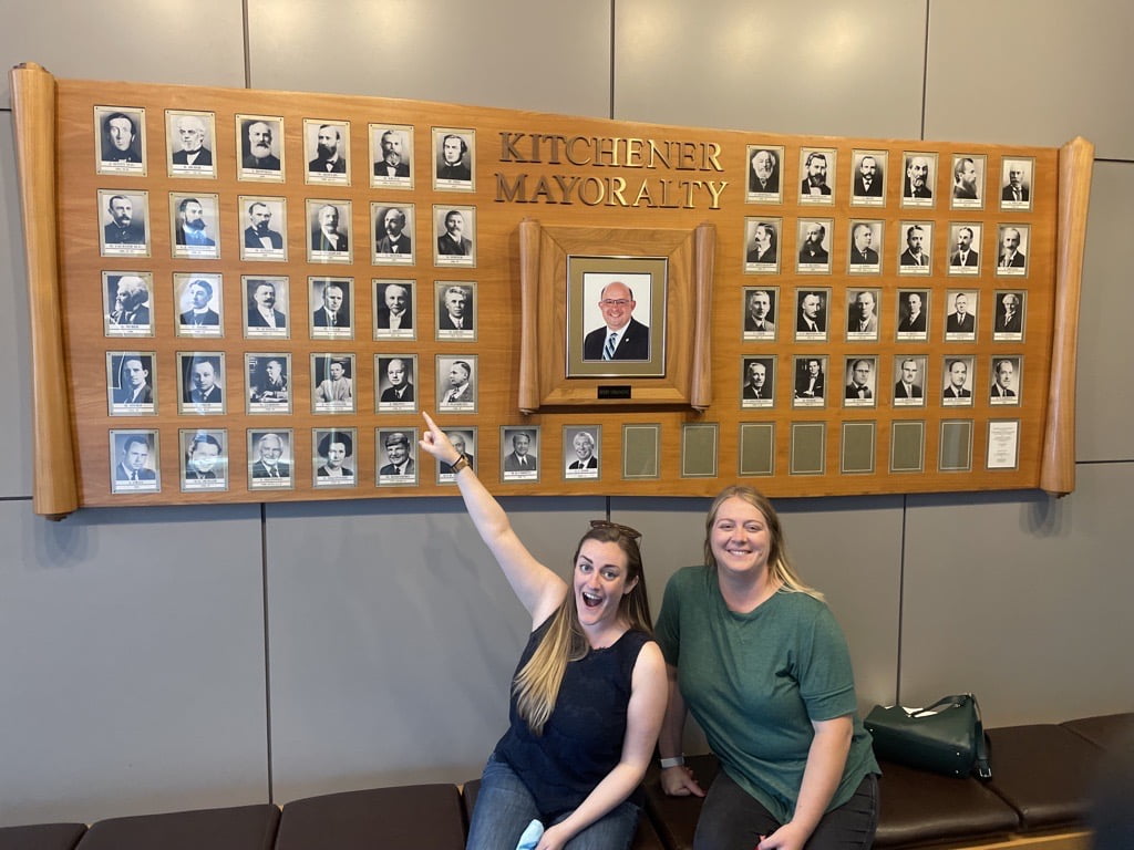 Chelsea and Joanna in front of Kitchener's Wall of Mayoralty and point towards J.M. Staebler's portrait